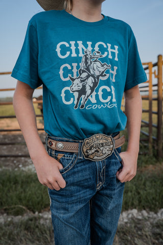 Child wearing a teal 'Cinch' branded t-shirt with a graphic of a person riding a bull, standing in an outdoor setting.