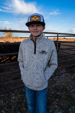Person wearing a gray pullover with a logo and a cap, standing in an outdoor setting with a fence and sky in the background.