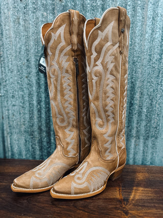Pair of tan cowboy boots with white embroidery on a wooden surface and blue corrugated metal background.