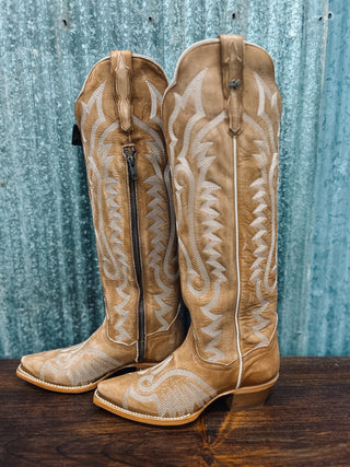 Pair of tan cowboy boots with intricate designs on a wooden surface and metal wall background