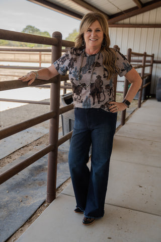 Woman standing in a barn-like setting wearing a patterned shirt and jeans.
