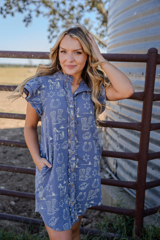 Woman in a blue dress with cowboy pattern standing in front of a metal silo.
