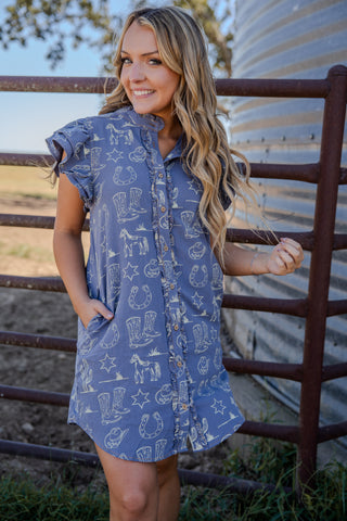Woman wearing a blue dress with cowboy print standing next to a metal fence.