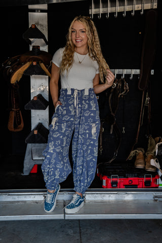 Woman wearing a white t-shirt and blue patterned pants standing in front of a dark background with equipment.