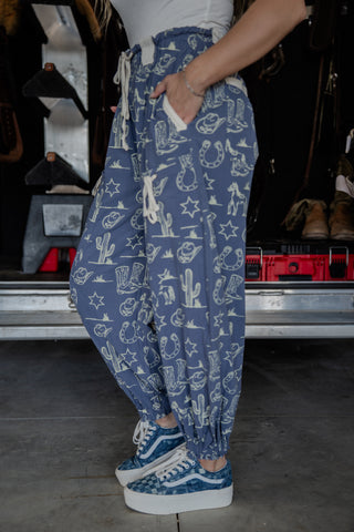 Person wearing blue patterned pants with a white shirt and blue sneakers in front of an open car trunk.