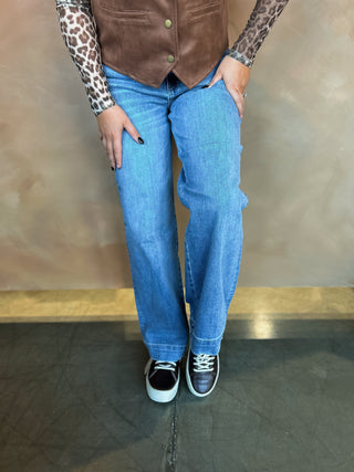 Person wearing a brown vest, blue jeans, and black sneakers against a neutral background