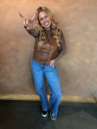Woman in a brown leather vest and blue jeans posing against a beige wall.