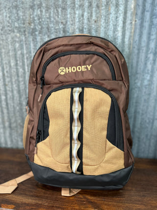 Brown and beige backpack with 'HOOey' logo on a wooden surface and metal wall background