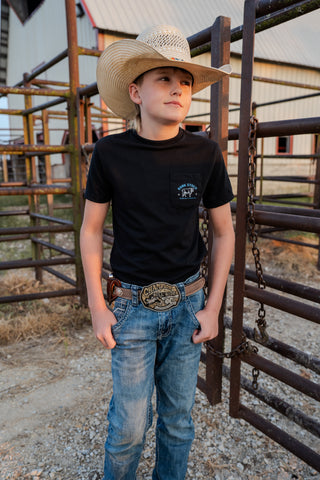 Young boy in a cowboy hat and black t-shirt standing in a rustic outdoor setting.