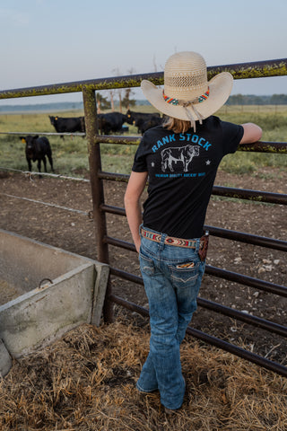Person wearing a cowboy hat and black t-shirt with a graphic design, standing in a fenced area with cows.