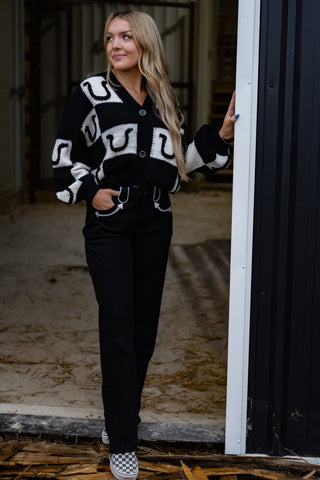 Woman wearing a black and white patterned cardigan and black pants, standing in a doorway.