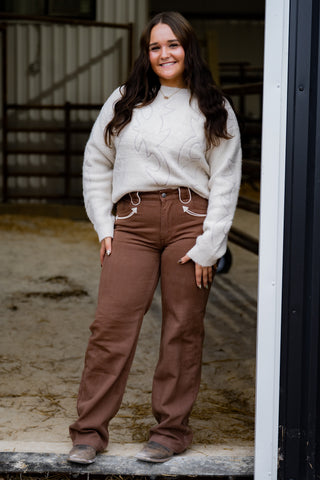Woman wearing a white sweater and brown pants standing in an outdoor setting.