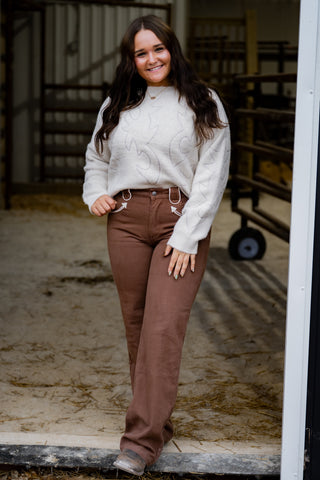 Woman wearing a white sweater and brown pants standing in an indoor setting.
