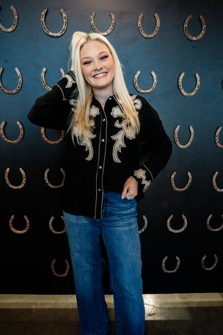 Person wearing a black embroidered shirt and blue jeans standing against a wall with horseshoe patterns.