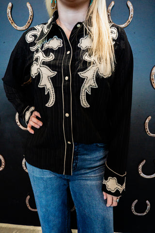 Person wearing a black embroidered shirt with white designs against a dark background with horseshoe patterns.