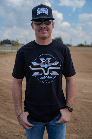 Man wearing a black t-shirt with a logo and a cap, standing on a dirt field.