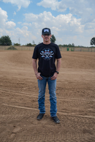 Man wearing a black t-shirt with a logo and blue jeans standing on a dirt field with trees and sky in the background.