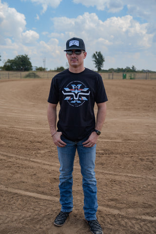 Man wearing a black t-shirt with a graphic design, blue jeans, and a cap on a dirt field.