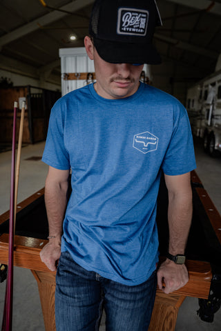 Man wearing a blue t-shirt with a logo and a black cap, standing in a garage.