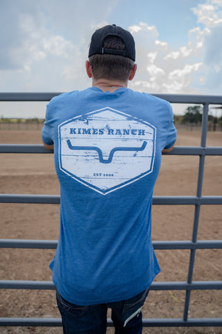 Person wearing a blue t-shirt with 'Kimes Ranch' logo standing in front of a fence.