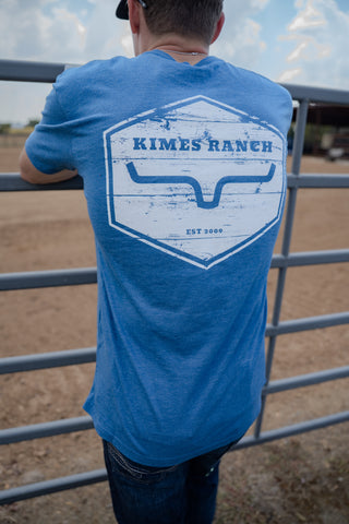 Person wearing a blue 'Kimes Ranch' t-shirt standing in front of a metal gate.