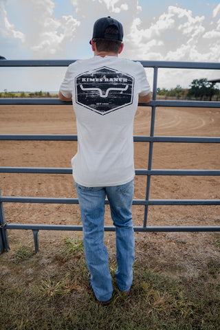 Person wearing a white t-shirt with a logo, standing in front of a metal fence with a rural landscape in the background.