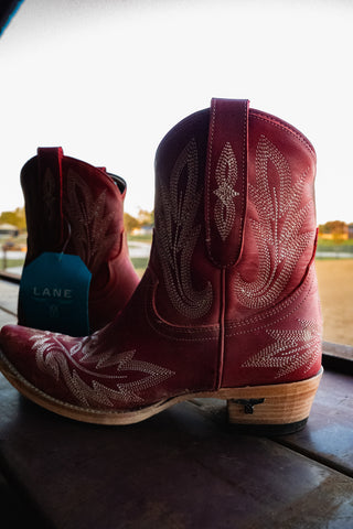 Red cowboy boots with intricate designs on a wooden surface, with a blurred outdoor background.