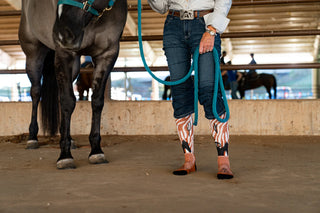 Person standing next to a horse in an indoor arena, wearing patterned cowboy boots.