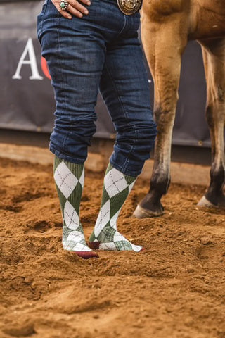 Person wearing argyle socks and riding pants standing next to a horse on a dirt ground.