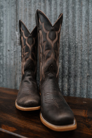 Pair of black cowboy boots with brown stitching on a wooden surface and metal background