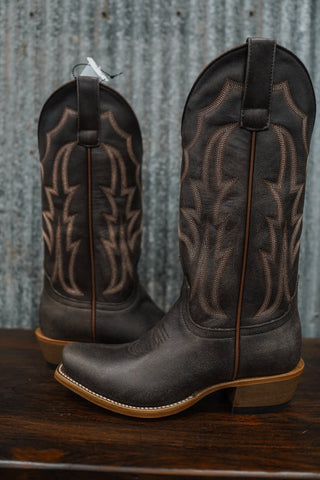 Pair of brown cowboy boots with intricate patterns on a wooden surface and metal wall background.