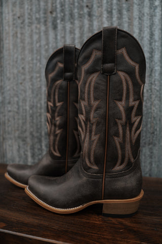 Pair of black cowboy boots with intricate designs on a wooden surface and corrugated metal background.