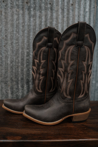 Pair of black cowboy boots with brown leather patterns on a wooden surface and metal wall background.