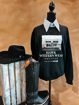 Black sweatshirt with 'Hawk Western Wear' logo on a mannequin, accompanied by a cowboy hat and boots against a brown background.