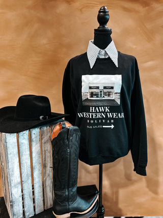 Black sweatshirt with graphic design on a mannequin, next to a cowboy hat and boots against a wooden background
