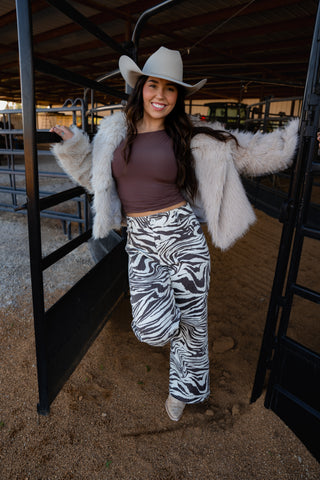 Woman in a barn wearing a white cowboy hat, brown top, zebra print pants, and a white fur coat.