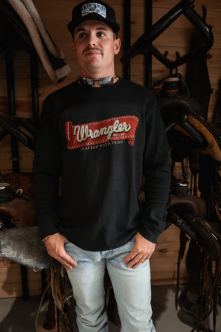 Man wearing a black Wrangler sweatshirt in a rustic setting with wooden walls and horse equipment.
