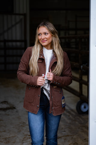 Woman wearing a brown jacket and blue jeans standing in an indoor setting.