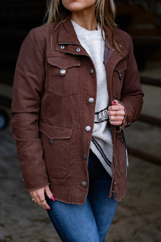 Person wearing a brown jacket over a white sweater with text, standing outdoors.