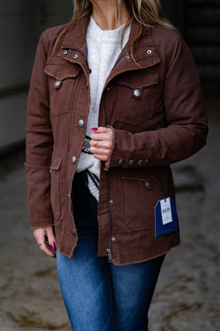 Person wearing a brown jacket with a visible brand tag, standing on a beach.
