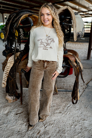 Woman standing in a stable wearing a light gray sweatshirt with a graphic design and brown pants.