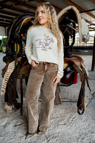 Woman wearing a white sweatshirt with a graphic design and brown pants standing in a stable with horse equipment.