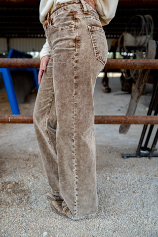 Beige corduroy pants worn by a person in an outdoor setting with a metal railing and blue chair in the background.