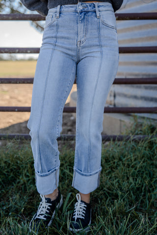 Person wearing light blue jeans and black sneakers standing in a grassy area with a metal fence in the background.