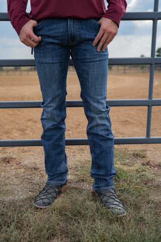 Person wearing blue jeans and a maroon shirt standing in front of a metal fence with a field in the background.