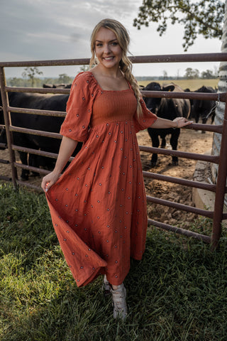 Woman in an orange dress standing next to a fence with cows in the background