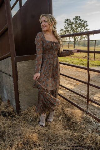 Woman in a patterned dress standing near a metal gate with a rural landscape in the background