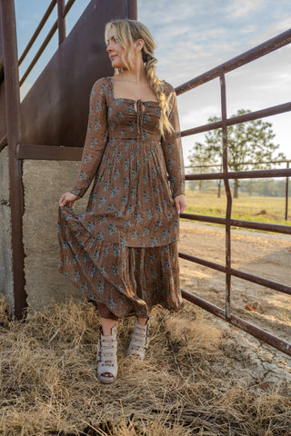 Woman in a brown floral dress standing in a rustic outdoor setting with a metal gate and field in the background.