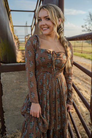 Woman in a brown floral dress standing in front of a metal fence with a rural background.