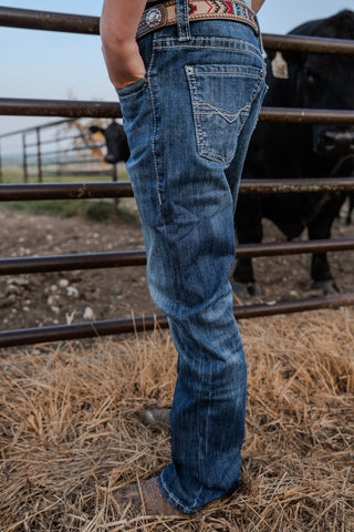Person wearing blue jeans standing near a metal fence with cattle in the background